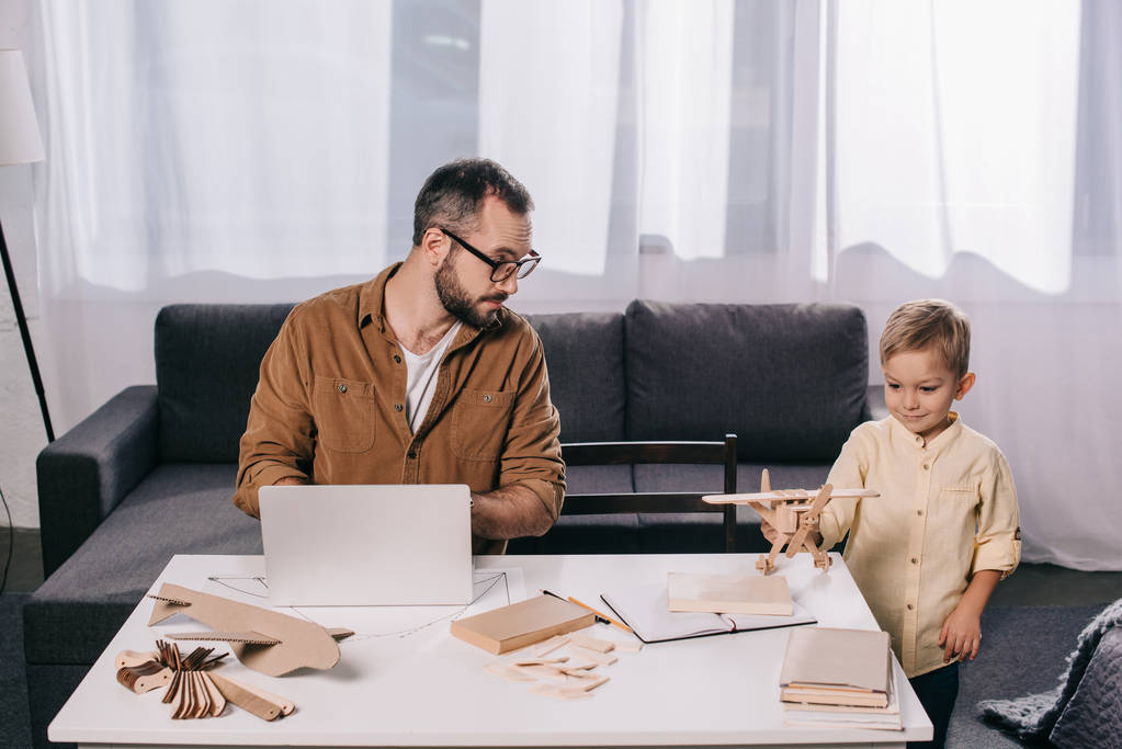 pai usando laptop e olhando para o pequeno filho brincando com modelo de avião de madeira em casa
 - Foto, Imagem