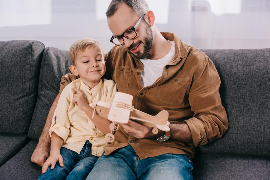Šťastný otec a roztomilý malý syn sedí na gauči a hrát si s dřevěné letadlo model    - Fotografie, Obrázek