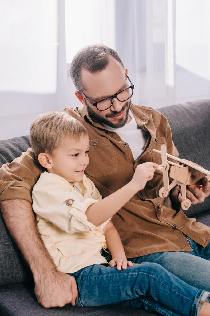 glücklicher Vater und Sohn sitzen auf der Couch und spielen mit einem hölzernen Flugzeugmodell   - Foto, Bild