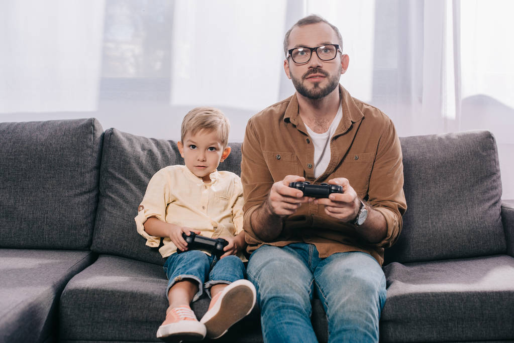 pai e pequeno filho sentado no sofá e brincando com joysticks
 - Foto, Imagem
