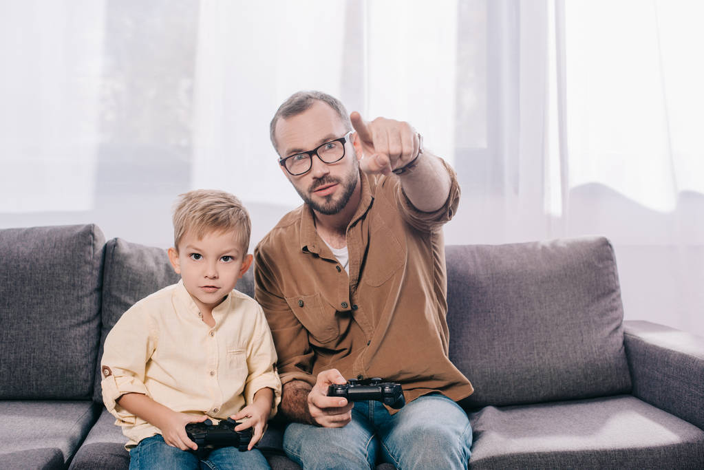 otec a malý syn sedí na gauči a hrát si s gamepady  - Fotografie, Obrázek