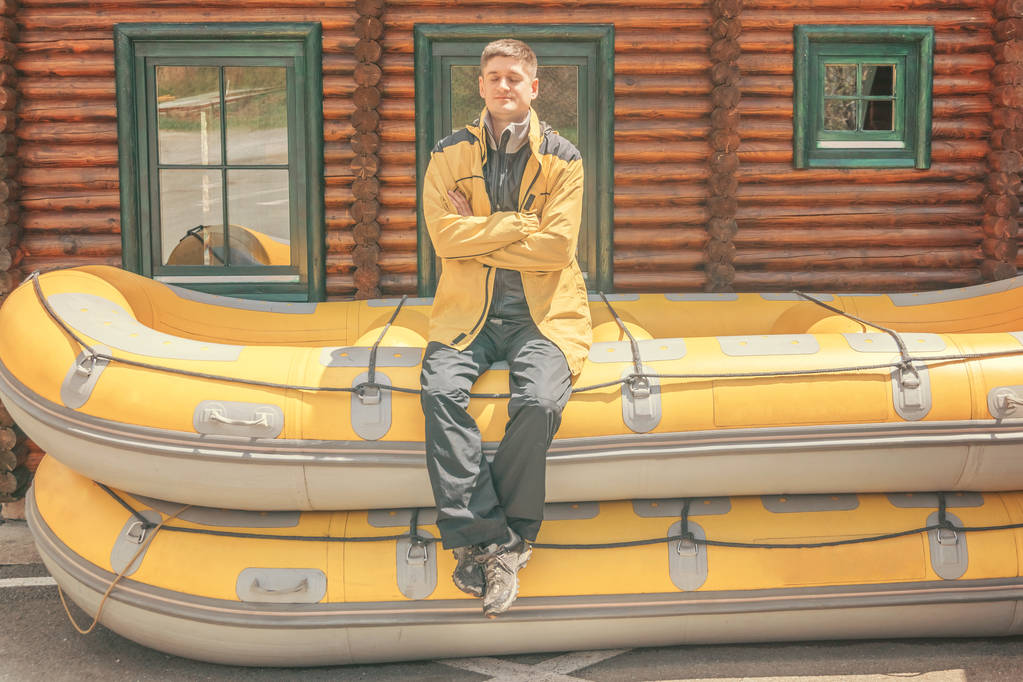 Dreamy muž ve žlutém kabátku, který sedí na dvou gumových lodích nedaleko lodního domu za slunného dne - Fotografie, Obrázek