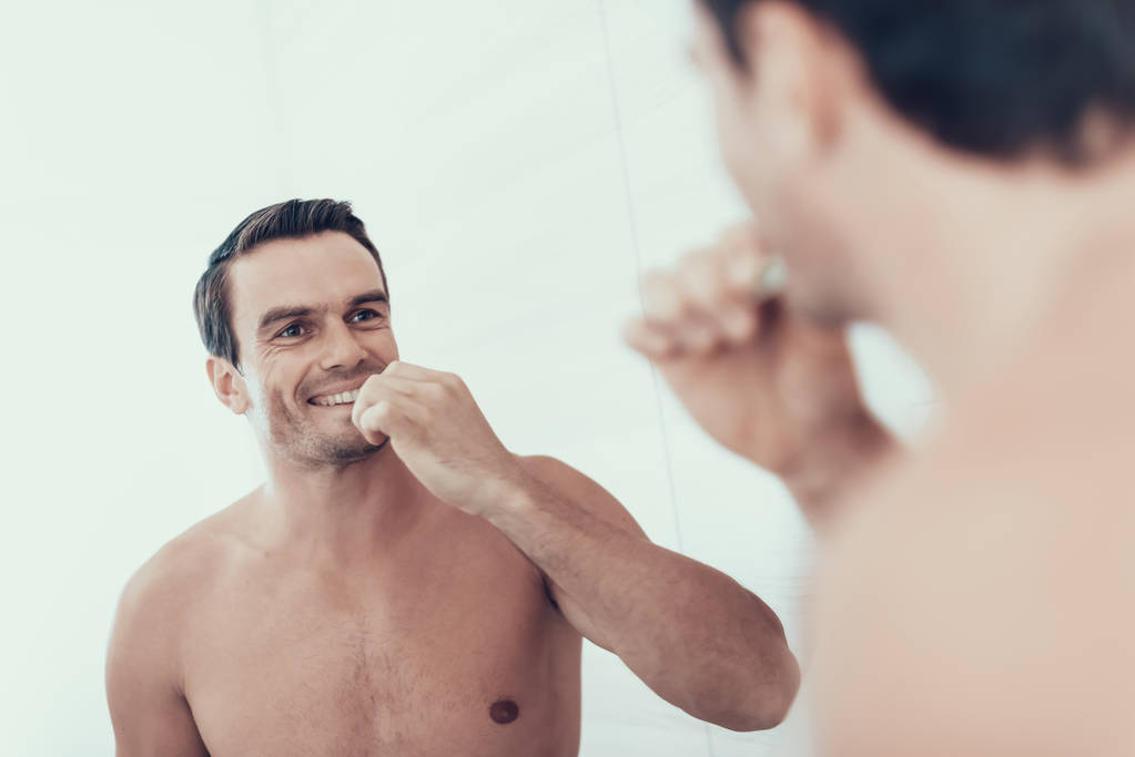 Espelho Reflexão dos dentes escova do homem no banheiro. Retrato de sorriso bonito Brown Cabelo Jovem com peito nu Segurando escova de dentes e olhando para o espelho. Conceito da manhã
 - Foto, Imagem