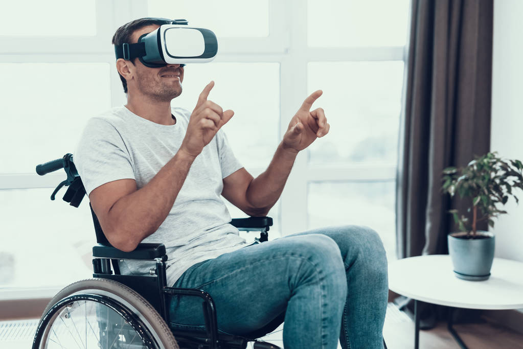 El hombre discapacitado disfruta de las gafas VR con las manos arriba. Retrato de una persona discapacitada sentada en silla de ruedas estacionada en una gran sala de estar luminosa. Hombre en ropa casual señalando dedo en la ventana
 - Foto, Imagen