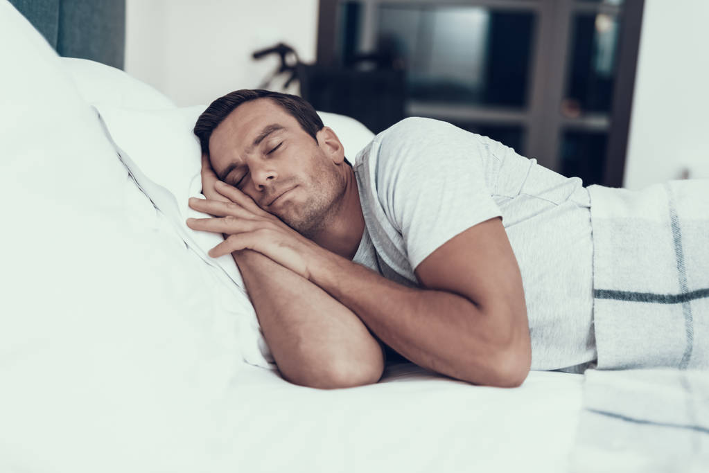 Person schläft in der Nähe von Alarm im Bett mit weißer Bettwäsche. schöner junger Mann, der mit verschwommenem Rollstuhl auf dem Rücken im Schlafzimmer liegt, die Hände unter dem Kopf hält und ein weißes T-Shirt trägt. Morgenkonzept - Foto, Bild
