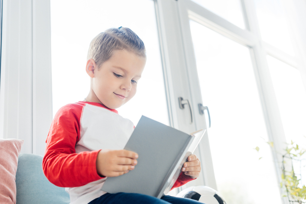 χαμογελώντας μικρό παιδί βιβλίο ανάγνωσης στο παράθυρο του σπιτιού - Φωτογραφία, εικόνα