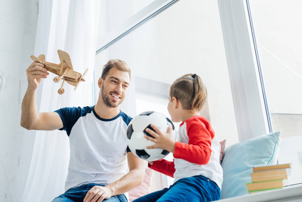 niño con pelota de fútbol mirando sonriente padre con avión de juguete de madera en casa
 - Foto, imagen