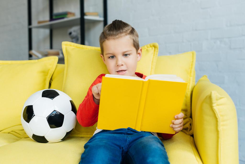πορτρέτο του μικρό αγόρι ανάγνωση βιβλίων στον κίτρινο καναπέ με ποδόσφαιρο μπάλα κοντά από στο σπίτι - Φωτογραφία, εικόνα