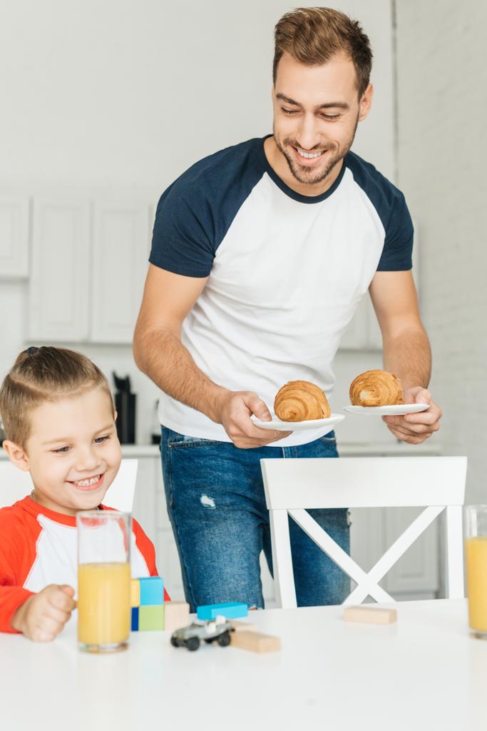 komea nuori isä tarjoilee croissanteja aamiaiseksi pojan kanssa leikkiessään leluilla
 - Valokuva, kuva