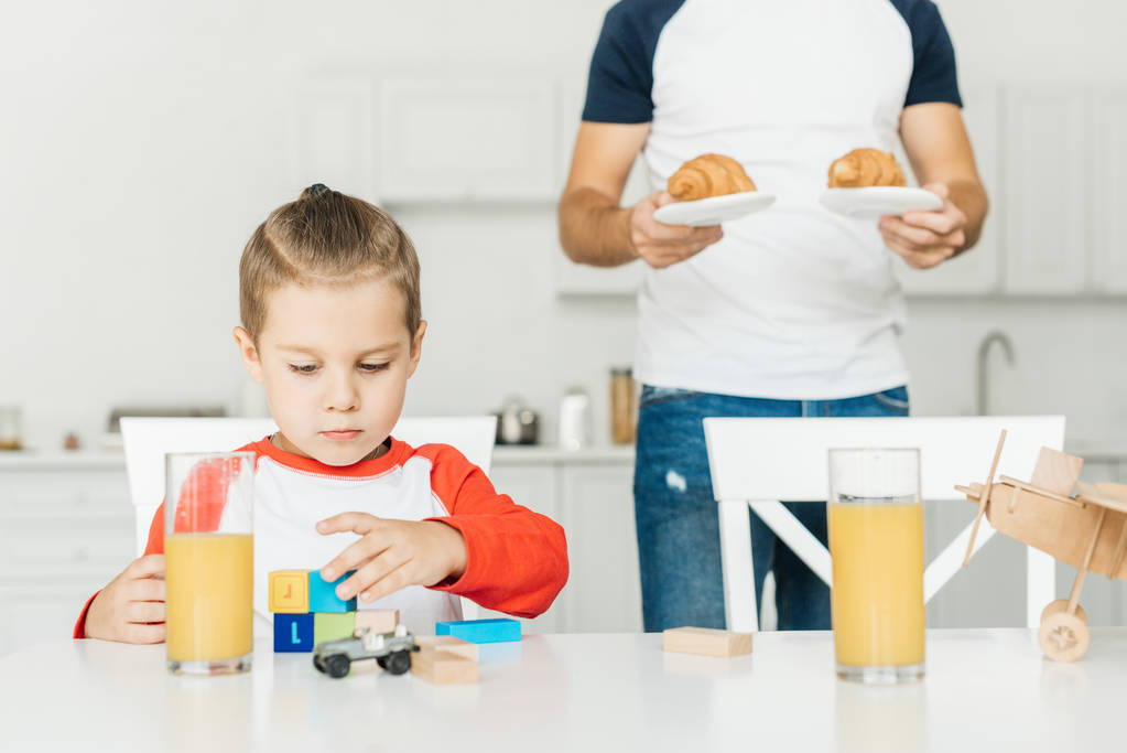 recortado tiro de padre sirviendo croissants para el desayuno con hijo mientras él jugando con juguetes
 - Foto, imagen
