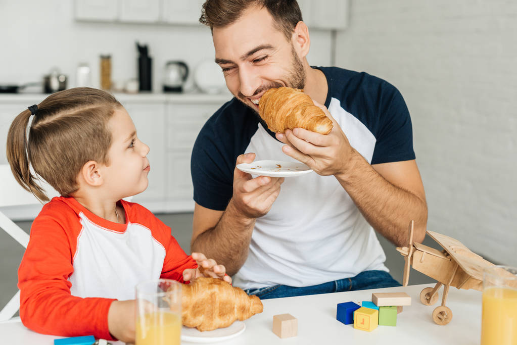 boldog fiatal apa és fiú birtoklás reggeli croissant-t és a narancslét együtt otthon - Fotó, kép