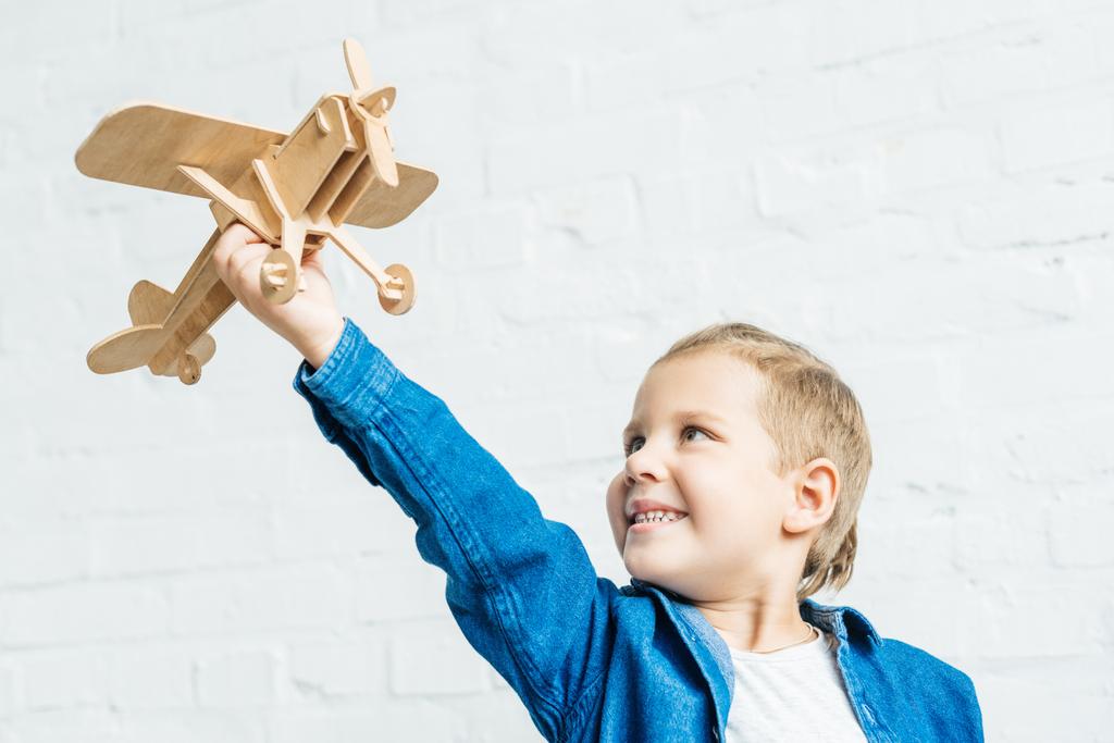 glimlachend klein kind spelen met houten speelgoed vliegtuig voor witte bakstenen muur - Foto, afbeelding