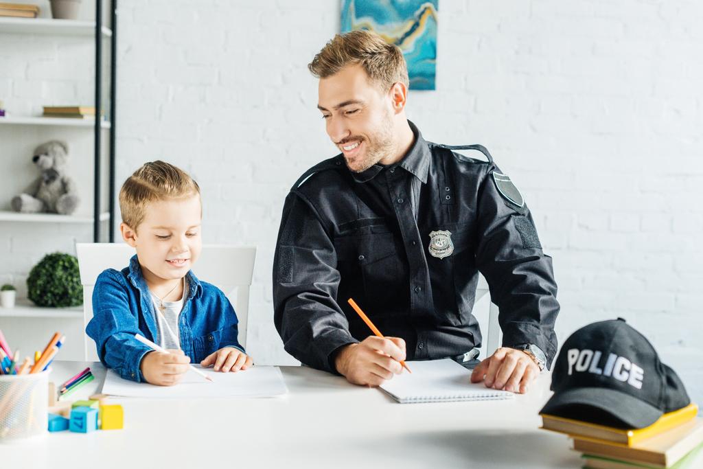 Улыбающийся молодой отец в полицейской форме и сын рисуют вместе дома
 - Фото, изображение