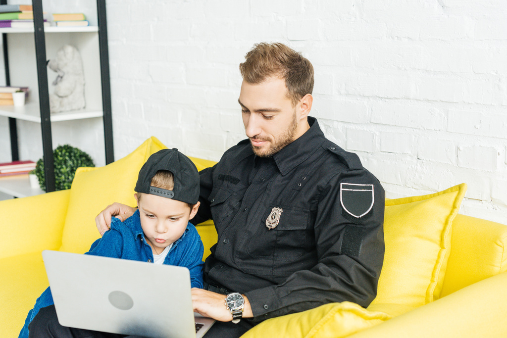 pohledný mladý otec v policejní uniformě a syn společně pomocí notebooku zatímco sedí na žluté pohovce doma - Fotografie, Obrázek