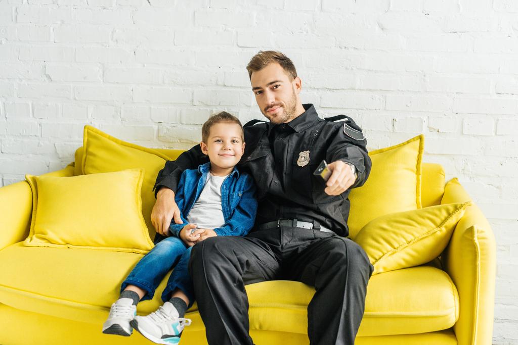 красивый молодой отец в полицейской форме смотрит телевизор с сыном, сидя дома на жёлтом диване
 - Фото, изображение