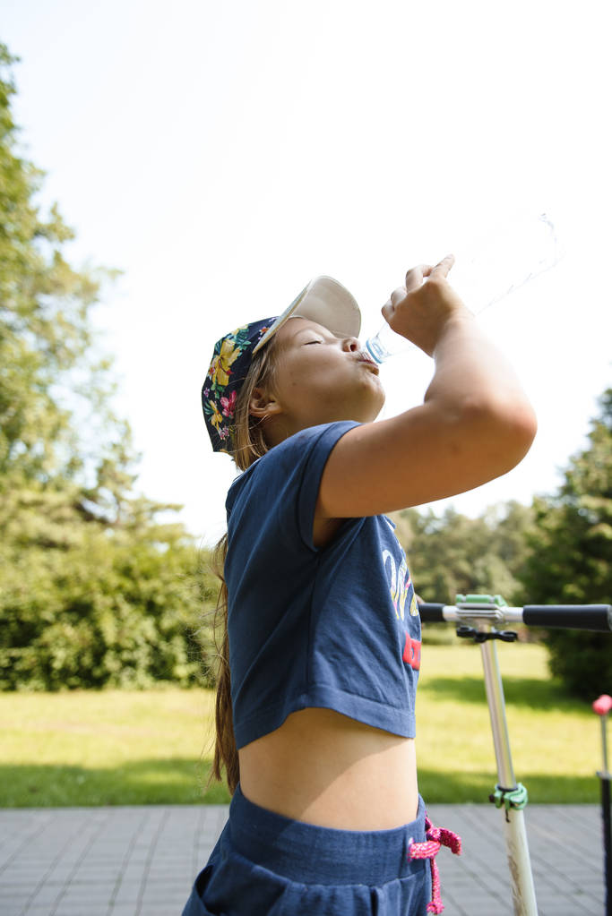 aranyos lány kék ruha ivóvíz palackból, míg a robogó lovaglás a napfényes nyári parkban  - Fotó, kép