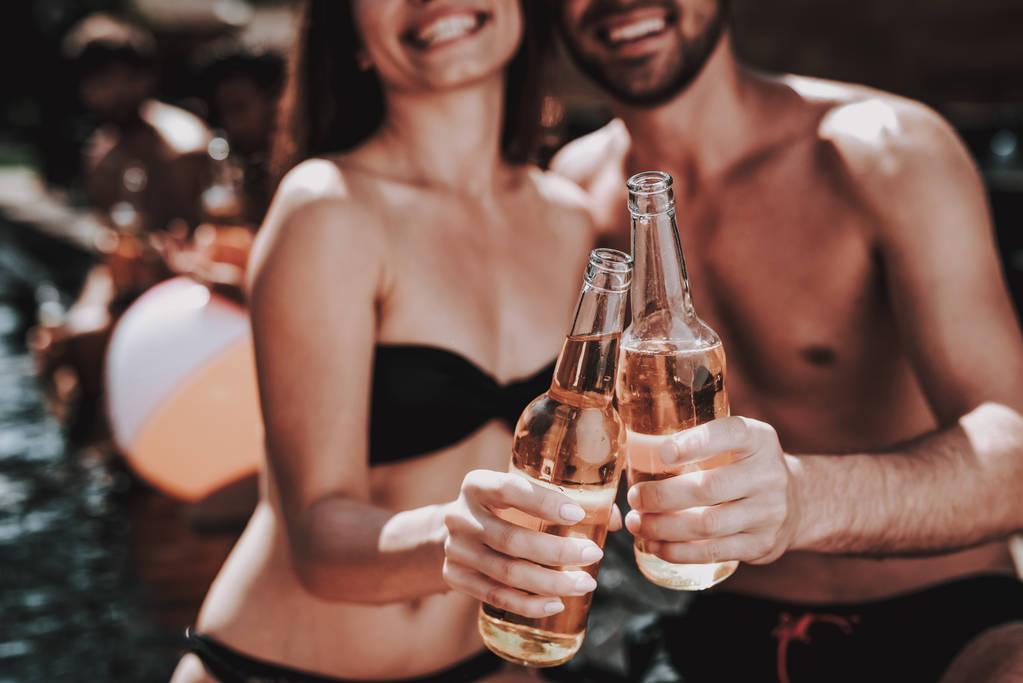 Alkoholos italokat a medence melletti házaspár mosolyog. Közelkép a gyönyörű fiatal pár gazdaság üveg sört és érzik magukat a medence mellett. Boldog meg Enoying Pool Party. Nyaralás koncepció - Fotó, kép