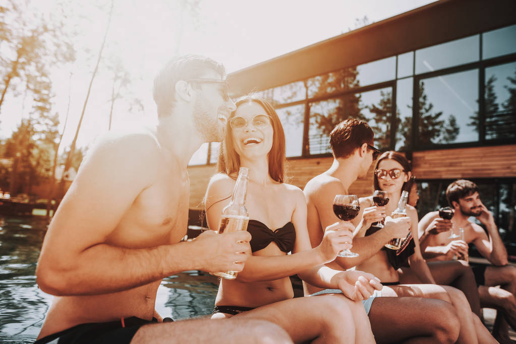 Nuoret hymyilevät ystävät juomassa viiniä Poolsidessa. Ryhmä nuoria onnellisia ihmisiä, jotka istuvat uima-altaalla yhdessä puhumassa ja pitämässä viinilaseja. Onnelliset ystävät uima-allasjuhlissa. Kesäloman käsite
 - Valokuva, kuva