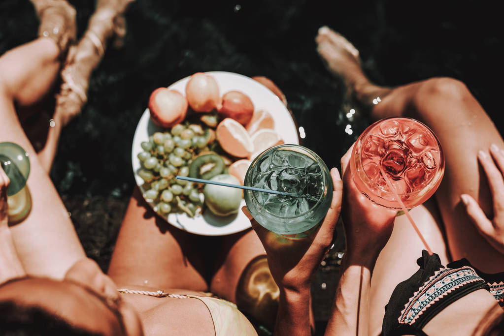 Jonge vrouwen met vruchten en Cocktails bij zwembad. Close-up van groep van jonge mooie vrouwen zitten bij zwembad holding kleurrijke alcoholische Cocktails en kom voor vers fruit. Zomervakantie Concept - Foto, afbeelding