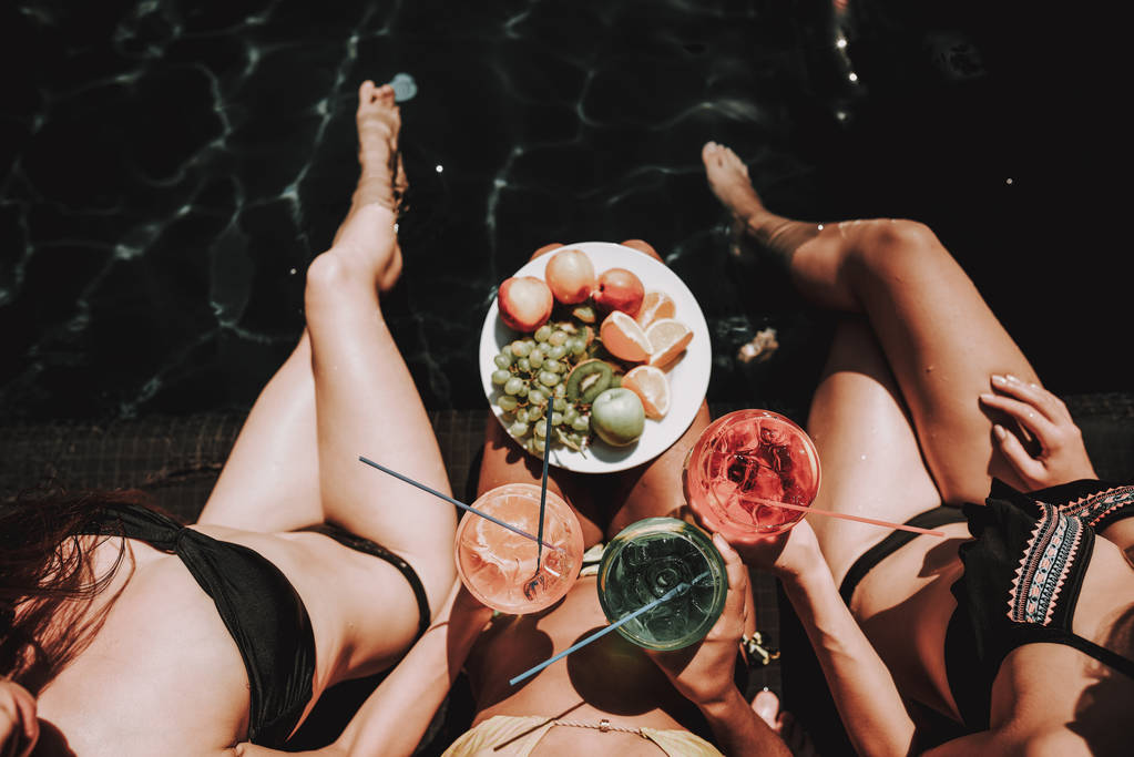 junge Frauen mit Früchten und Cocktails am Pool. Draufsicht auf eine Gruppe junger schöner Frauen, die am Pool sitzen und bunte alkoholische Cocktails und eine Schüssel mit frischem Obst in der Hand halten. Sommerferienkonzept - Foto, Bild