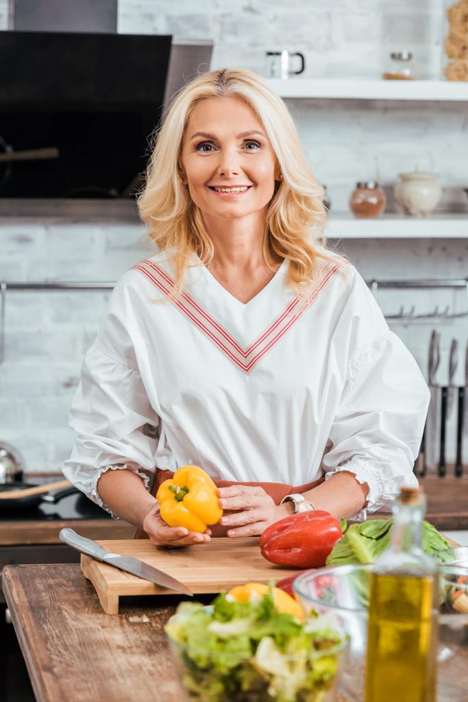 Улыбающаяся привлекательная женщина готовит салат к ужину, держит перец и смотрит в камеру дома
 - Фото, изображение