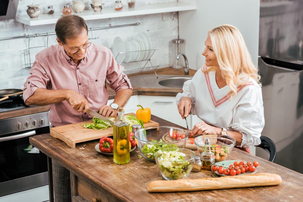 sorrindo marido e mulher adultos preparando salada para jantar juntos e cortar legumes na cozinha
 - Foto, Imagem