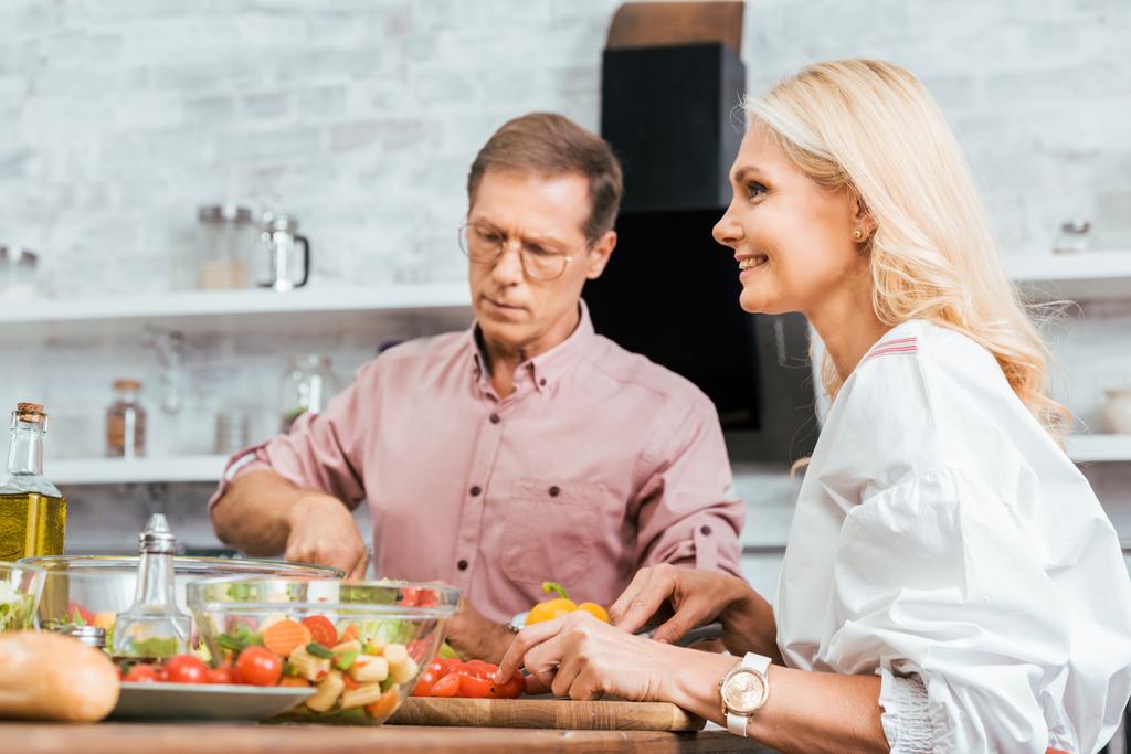 smiling adult couple preparing salad for dinner together in kitchen - Photo, Image