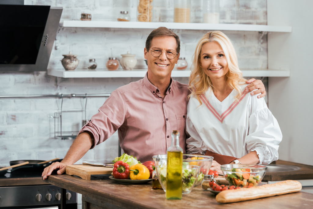 Erwachsenes Paar umarmt sich und blickt während der Salatzubereitung für das Abendessen zu Hause in die Kamera - Foto, Bild