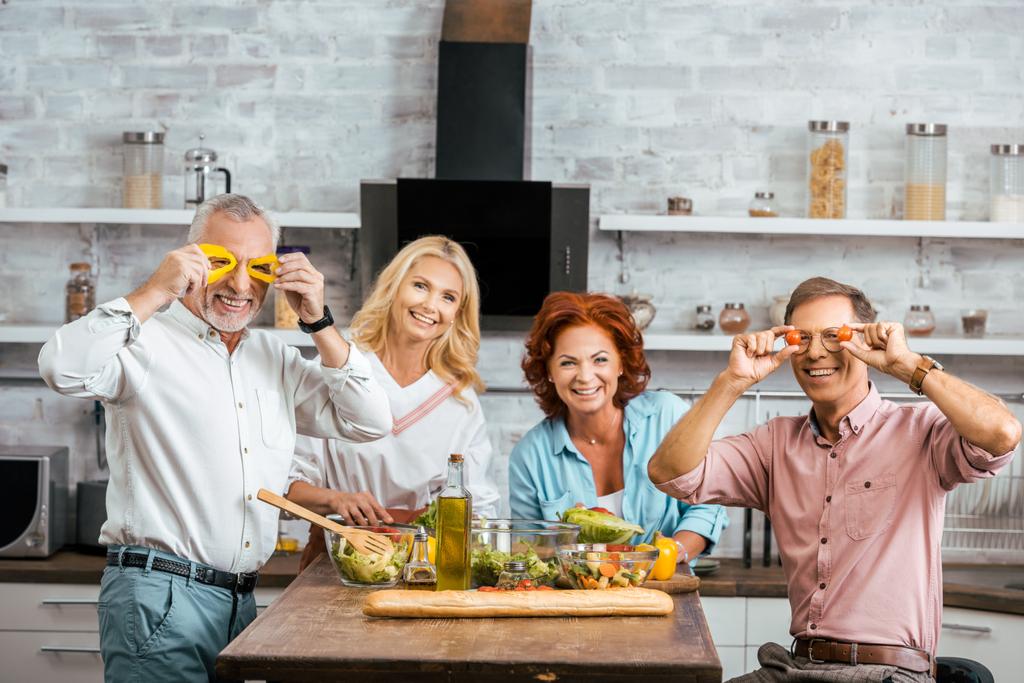 mature men having fun with vegetables during preparing salad for dinner at home and looking at camera - Photo, Image
