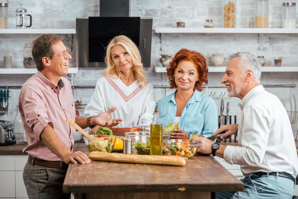 счастливые зрелые друзья готовят салат к ужину дома, мужчины разговаривают, женщины смотрят в камеру
 - Фото, изображение