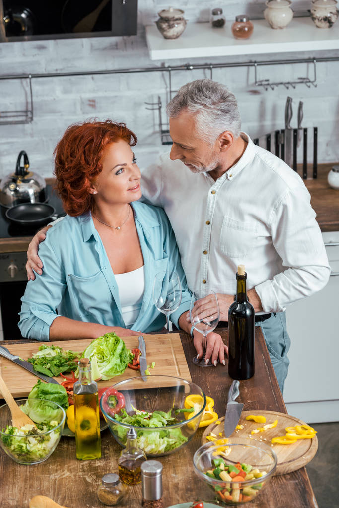 vista de ángulo alto del marido abrazando a la esposa durante la preparación de la ensalada para la cena y se miran entre sí
 - Foto, Imagen