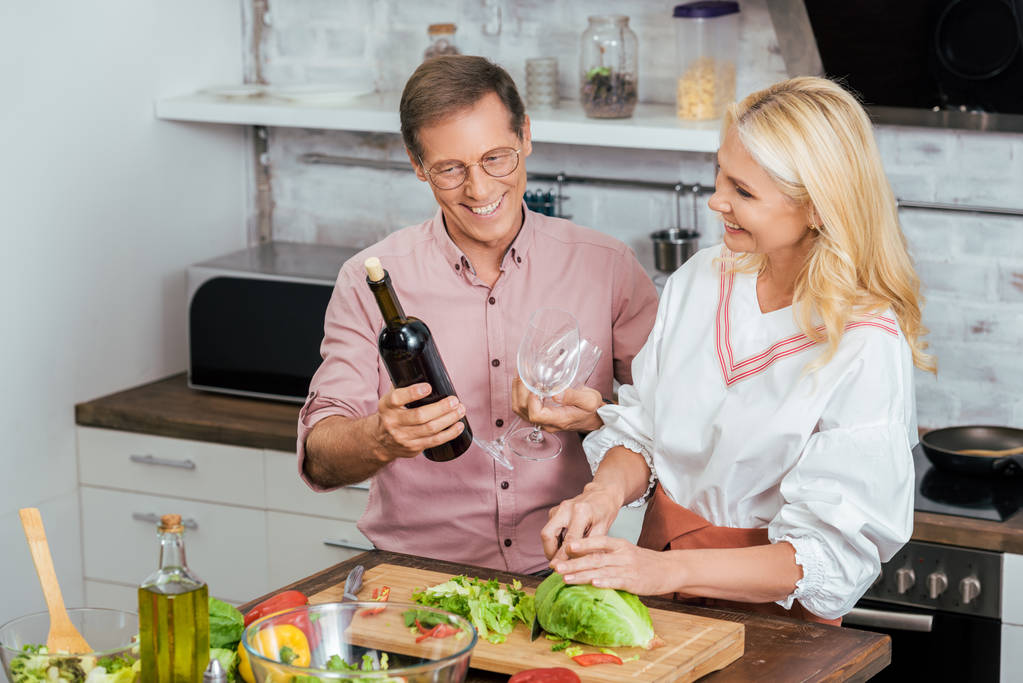 beautiful wife preparing salad for dinner at home, smiling husband holding wine bottle and wineglasses - Photo, Image