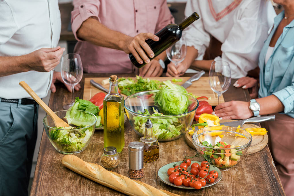 cropped image of man pouring wine to happy old friends during dinner in kitchen, vegetables on tabletop - Photo, Image