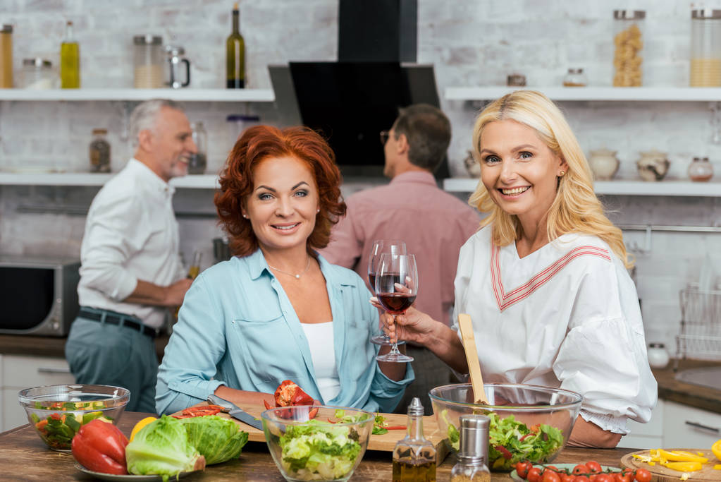 hymyilevät kauniit naiset valmistelevat salaattia päivälliselle, pitävät viinilaseja ja katsovat kameraa kotona
 - Valokuva, kuva