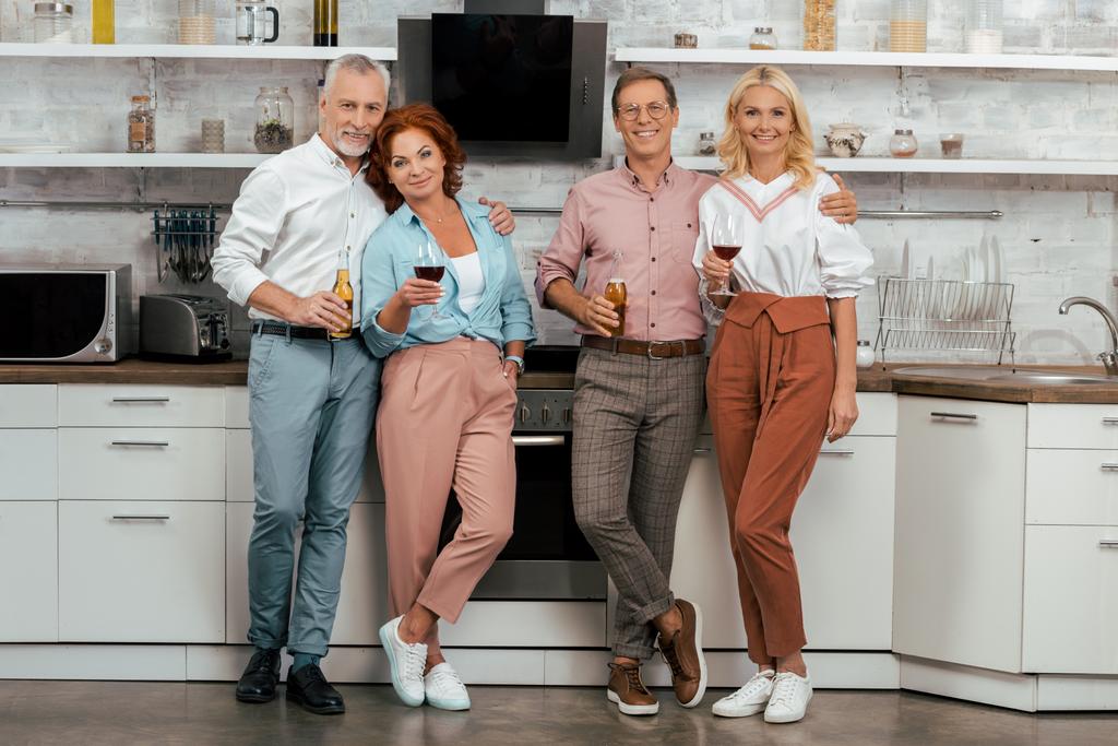 two happy mature couples holding alcoholic beverages and smiling at camera in kitchen  - Photo, Image