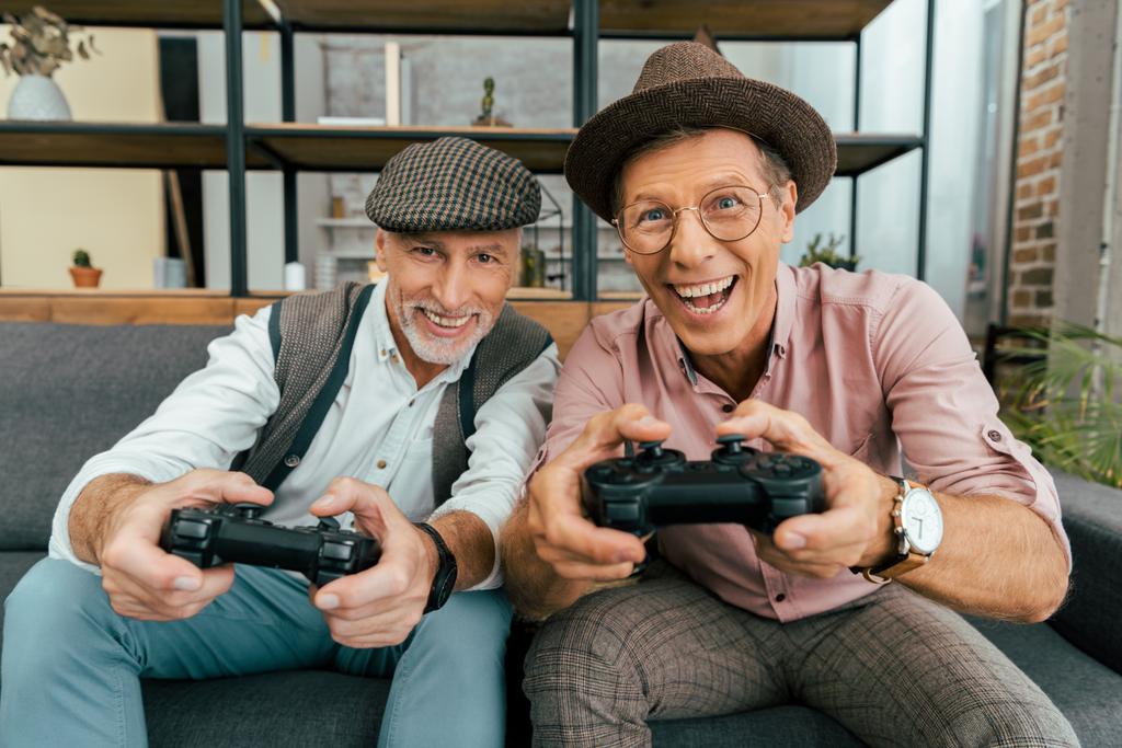hombres maduros emocionados jugando con joysticks y sonriendo a la cámara
 - Foto, Imagen