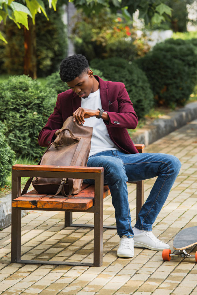 stylish young student with skateboard opening backpack while sitting on bench on street - Photo, Image