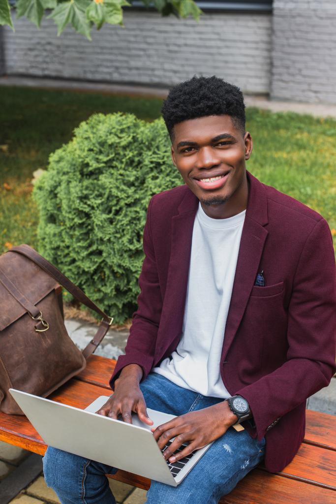 hymyilevä nuori afrikkalainen amerikkalainen opiskelija käyttää kannettavaa tietokonetta kadulla ja katselee kameraa
 - Valokuva, kuva