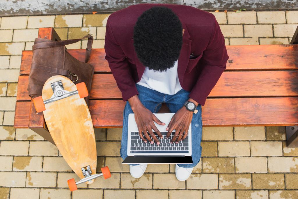 top view of young freelancer using laptop on bench with leather backpack and skateboard - Photo, Image
