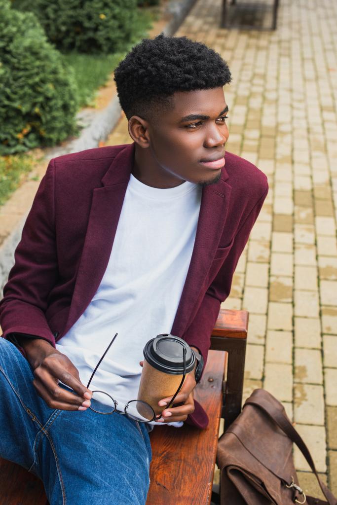 Красивый молодой человек с бумажной чашкой кофе и очками сидит на скамейке запасных
 - Фото, изображение
