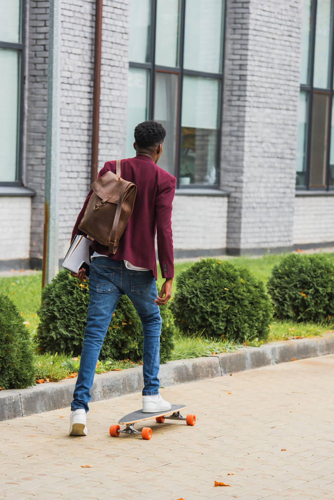rear view of stylish student with backpack and notebooks riding on skateboard on street - Photo, Image