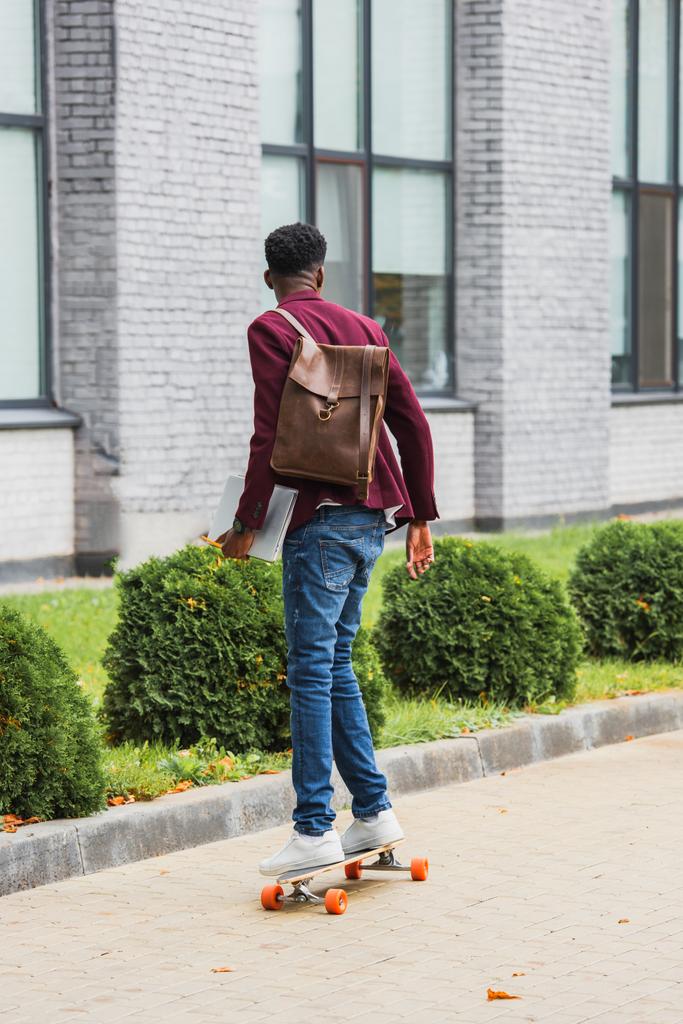 vue arrière de l'élève avec sac à dos et cahiers chevauchant sur planche à roulettes sur rue
 - Photo, image