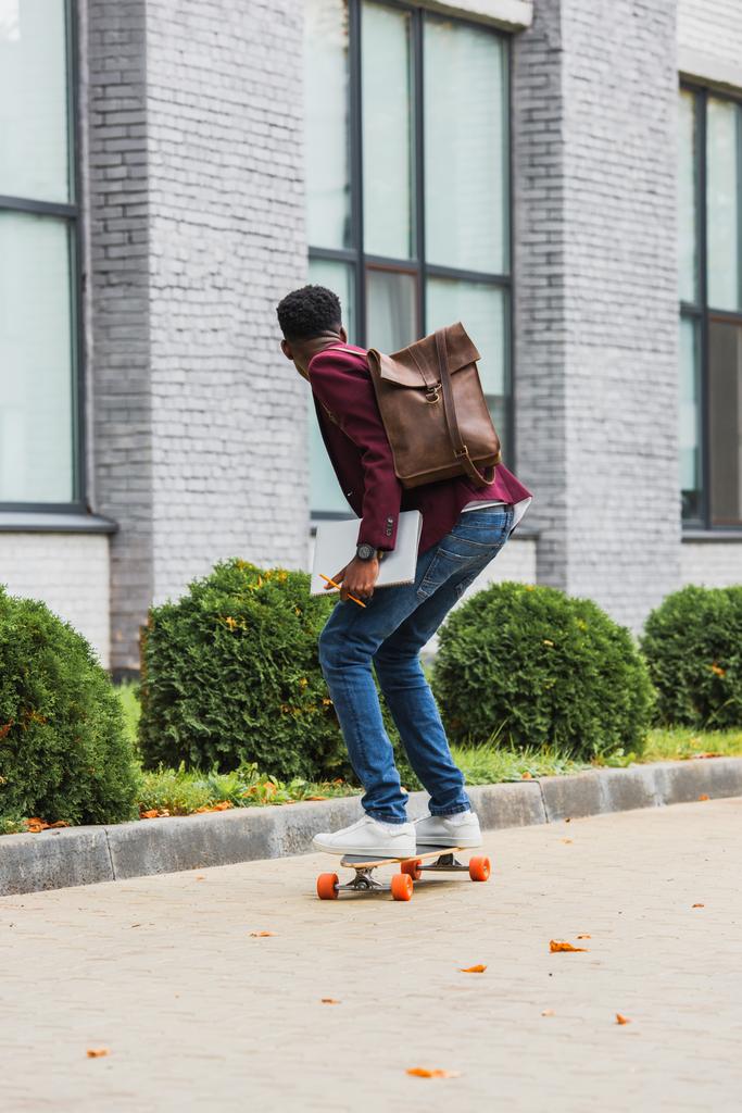 вид сзади на молодого студента с рюкзаком и ноутбуками, катающегося на скейтборде по улице
 - Фото, изображение