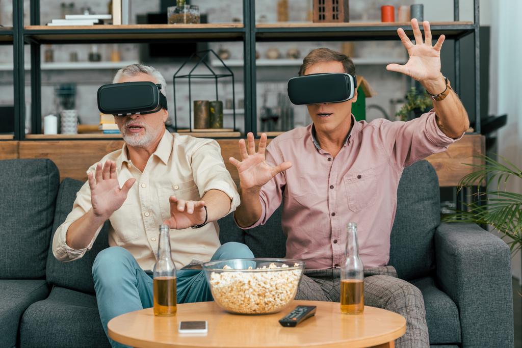 зрілі чоловіки друзі використовують гарнітури віртуальної реальності під час пиття пива та їжі попкорну вдома
 - Фото, зображення