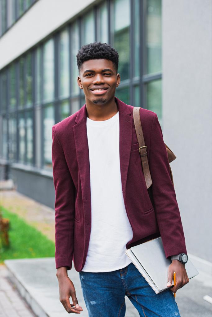 glücklicher junger Student mit Rucksack und Notizbüchern, der auf der Straße in die Kamera schaut - Foto, Bild