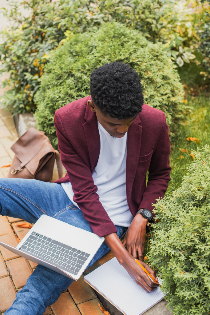 hoge hoekmening van jonge freelancer gebruik van laptop en het schrijven van notitieblok zittend op de stoeprand op straat - Foto, afbeelding
