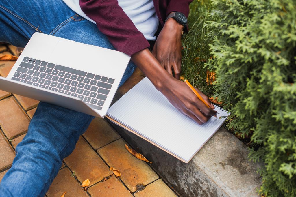 tiro recortado de freelancer usando laptop e escrever em notebook enquanto sentado no passeio na rua
 - Foto, Imagem