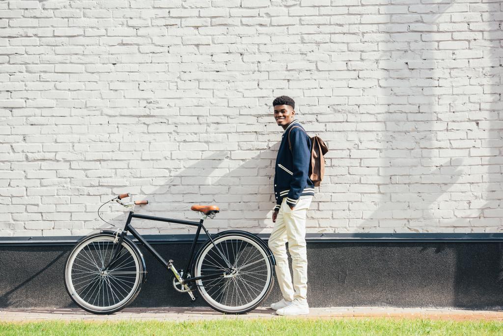 joyeux homme afro-américain élégant avec sac à dos en cuir debout avec vélo près du mur de briques blanches
 - Photo, image