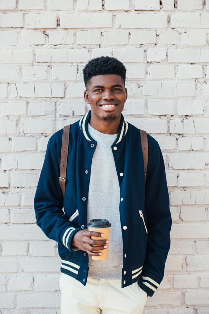 cheerful african american man holding coffee to go near white brick wall  - Photo, Image