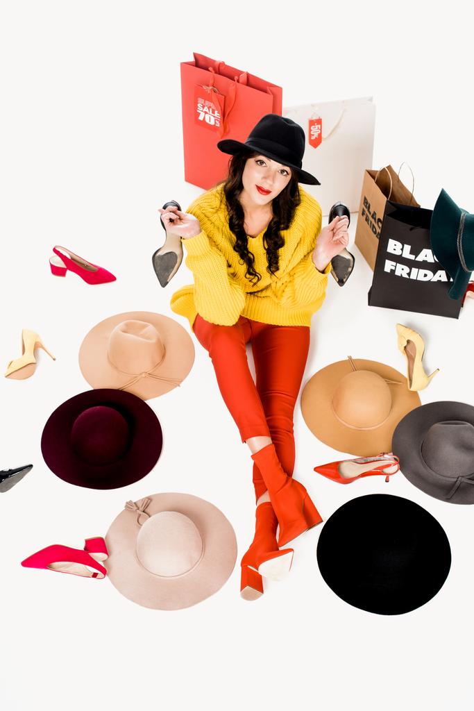 высокий угол зрения молодой женщины, сидящей на полу в обуви, шляпах и торговых мешках с черной пятничной надписью вокруг изолированных на белом
 - Фото, изображение